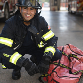 A photo of Rohan Shirur dressed in full firefighter gear, crouching down beside a duffle bag.