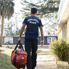 Image of Rohan Shirur walking away from the camera, wearing a black t shirt, dark pants, and carrying an orange duffle bag.